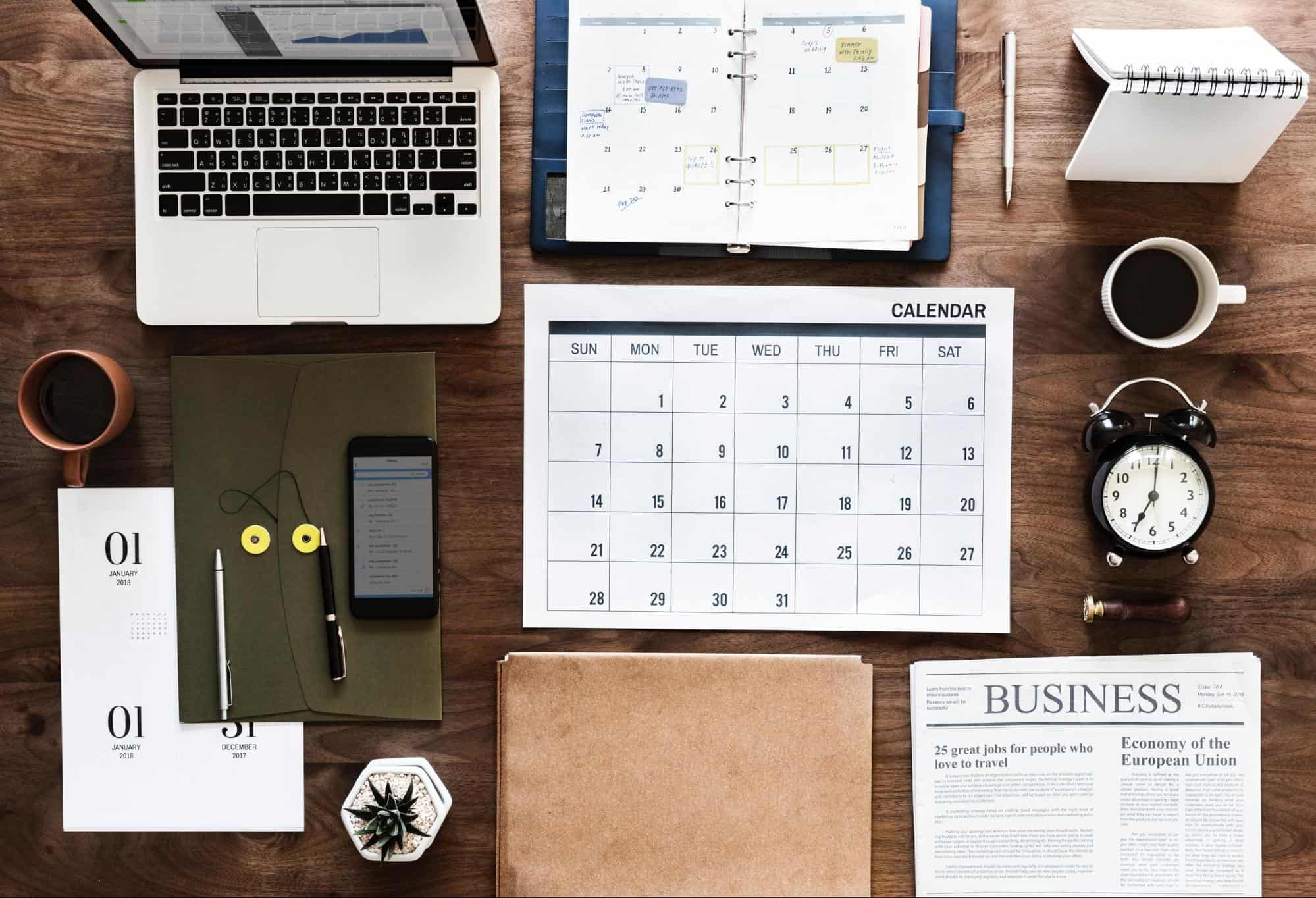 Calendar, alarm clock, planner, laptop on a desk.