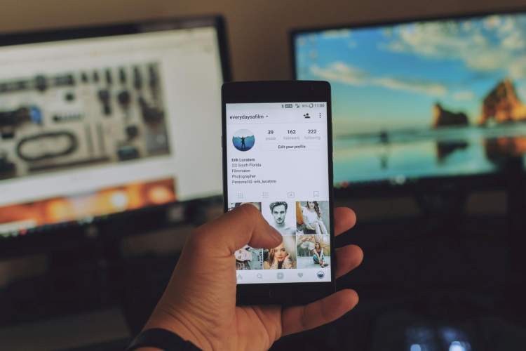 A hand holding a smart phone in front of two monitors.