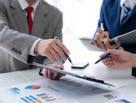 Three people in business attire discuss charts and graphs on a table. One person points to a document on a clipboard while another takes notes.