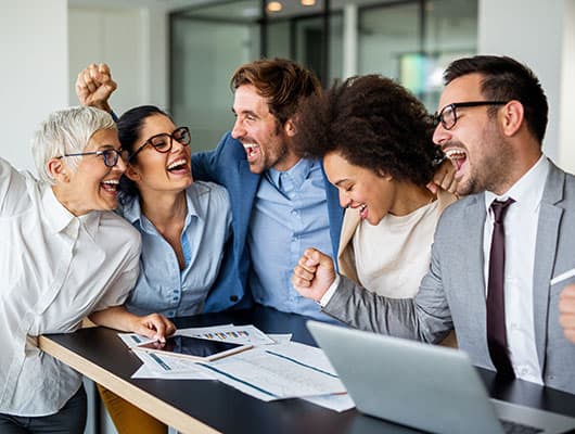 A group of five colleagues in an office celebrate a success, smiling and raising their fists around a desk with documents and a laptop.