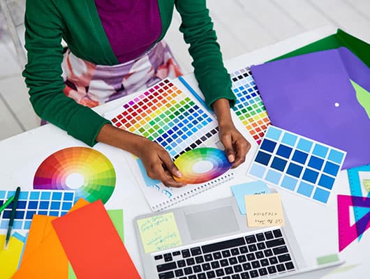 Person working at a desk with a laptop, color swatches, and various colored papers, seemingly involved in a design project.