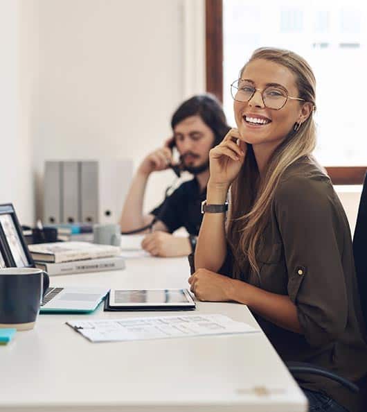 A woman in glasses is smiling at her desk with a tablet and papers. A man is working at a desk behind her, talking on the phone. Both are in a bright office space.