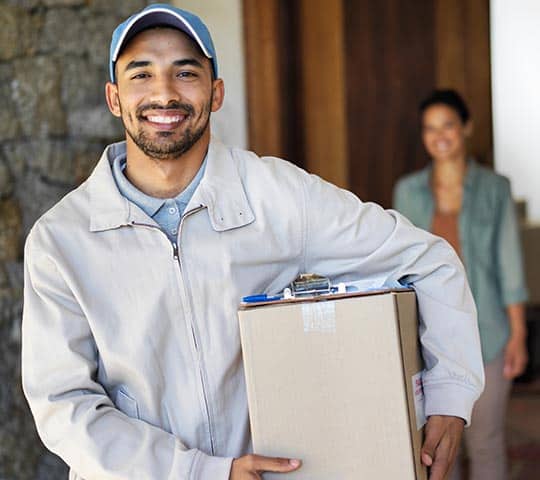 A delivery person wearing a cap and beige jacket smiles and holds a package, with a clipboard on top. A person in casual attire stands in the background near a doorway.