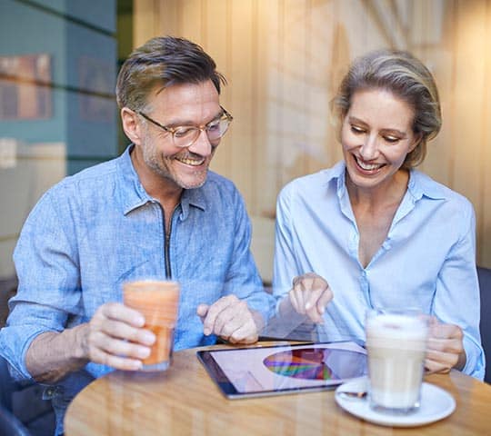 Two people sit at a café table, smiling and looking at a tablet. One holds a smoothie while the other points at the screen, which displays a colorful graph.