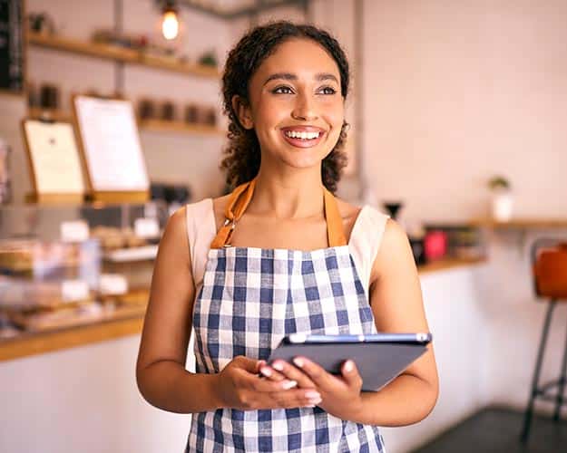 A smiling person wearing an apron holds a tablet in a cafe setting. Shelves, menu boards, and various items are visible in the background.