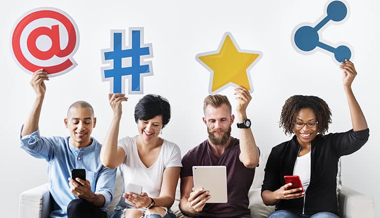 Four people sitting on a couch hold up large social media icons, including an at symbol, hashtag, star, and share symbol, while using electronic devices.