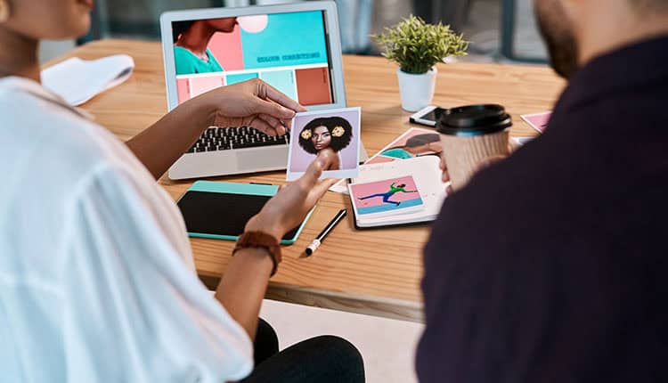 Two people review a photo at a desk with a laptop, a tablet, notebooks, and a cup of coffee, discussing creative design projects.