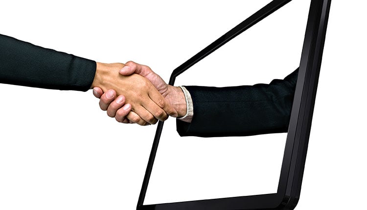 A hand reaches through a computer screen to shake hands with another person, symbolizing a virtual agreement or connection.