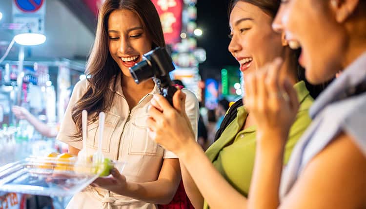 Three women smile while enjoying street food at night. One holds a camera, capturing the moment.