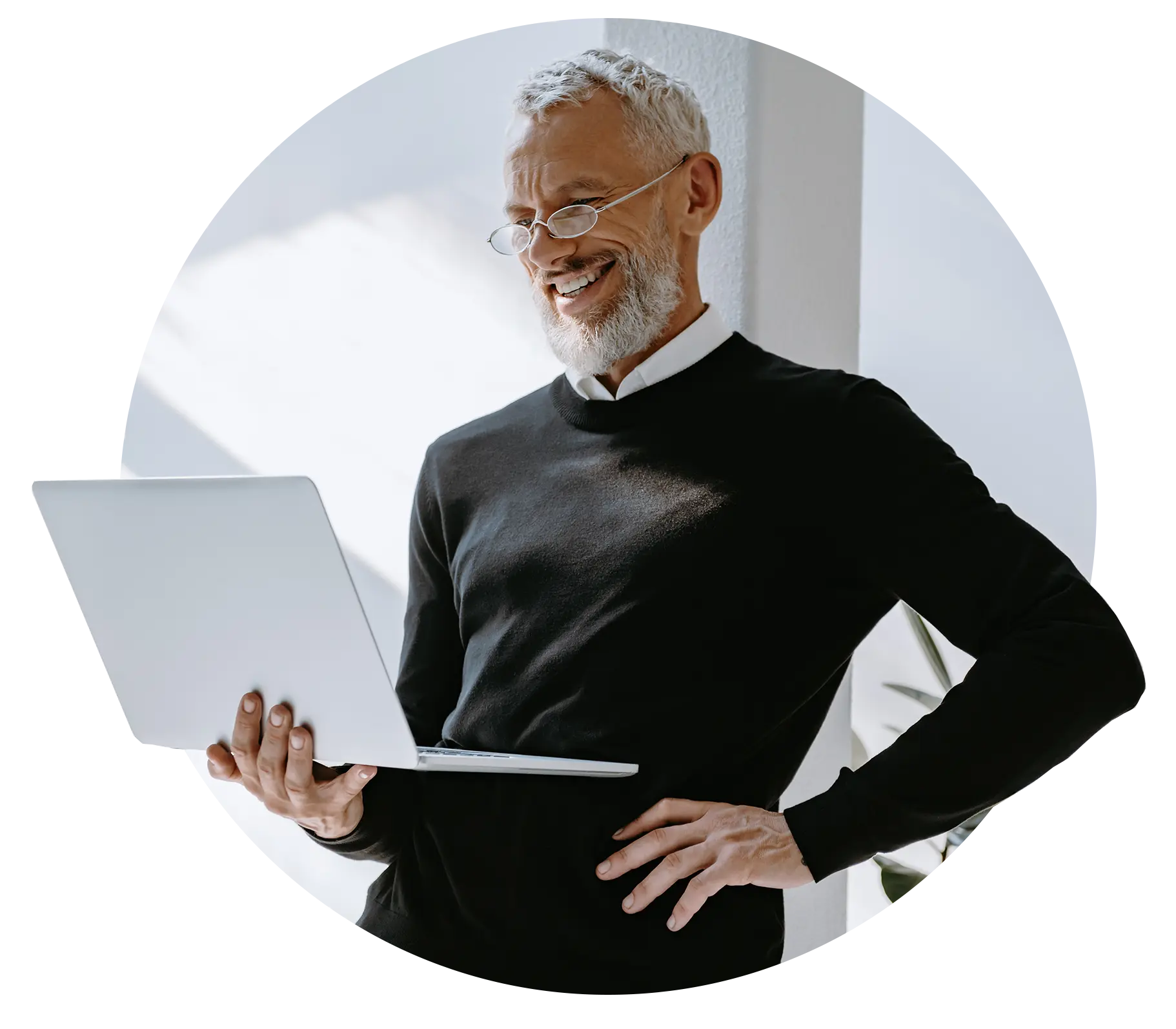 An older man with glasses smiles while looking at a laptop. He is standing near a window and wearing a black sweater over a white shirt.