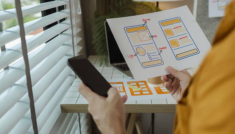 A person holds a smartphone in one hand and a mobile app wireframe sketch in the other, standing by a desk with design materials and a laptop.