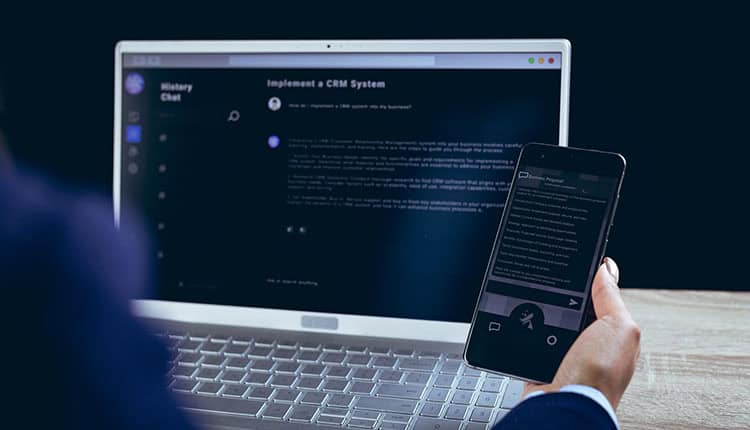 A person holds a smartphone displaying a chat while sitting in front of a laptop with a similar chat interface open on the screen.