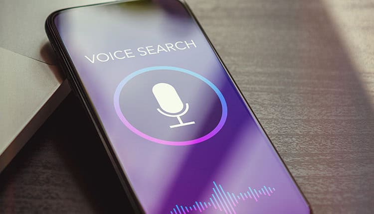 A smartphone screen displays a voice search interface with a microphone icon and sound wave graphic, resting on a table beside a laptop.
