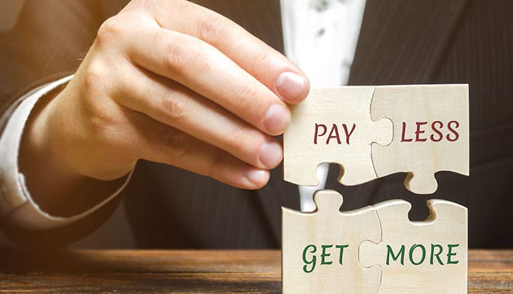 A person in a suit holds two wooden puzzle pieces that fit together, displaying the phrases "PAY LESS" and "GET MORE.