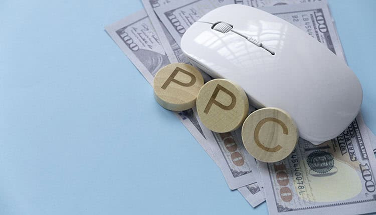 A computer mouse and three wooden blocks labeled "PPC" rest on top of several US hundred-dollar bills against a blue background.