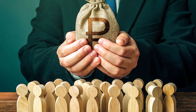 A person in a suit holds a sack with a currency symbol above a group of wooden human figures.
