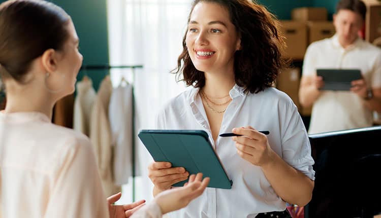Two women talk while one holds a tablet and stylus, smiling.