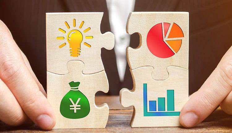 A person holds two puzzle pieces with symbols: a lightbulb, a pie chart, a money bag with a yen sign, and a bar graph, representing business concepts.
